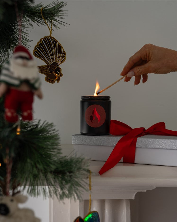 Person lighting a candle with a match near a Christmas tree and wrapped gift.